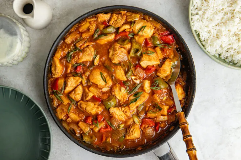 Overhead view of healthy sweet and sour chicken in a large frying pan, with chunks of chicken breast, red and green peppers, onion and pineapple coated in a glossy sweet and sour sauce. Served alongside fluffy white rice and finished with sliced spring onion.