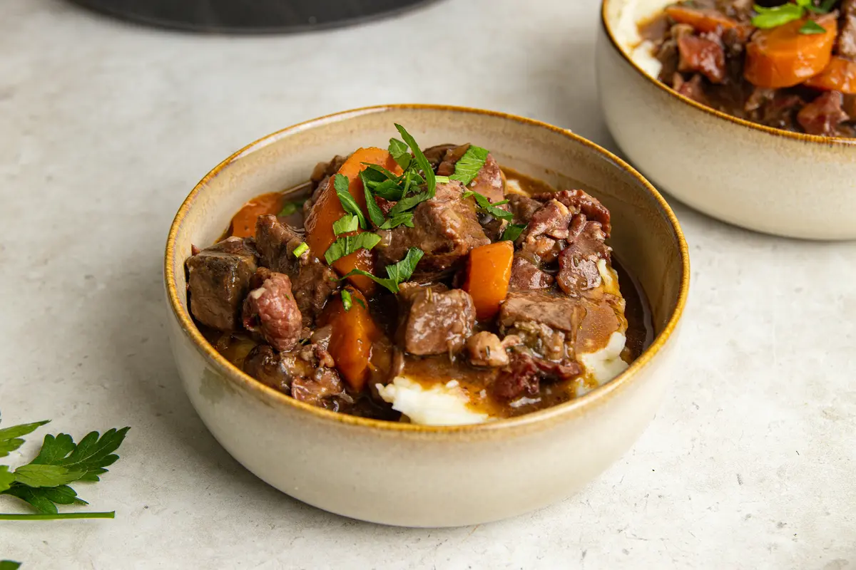 A comforting bowl of slow cooker beef bourguignon served over creamy mashed potato, with chunky beef, carrots and bacon coated in rich red wine gravy and topped with fresh parsley.