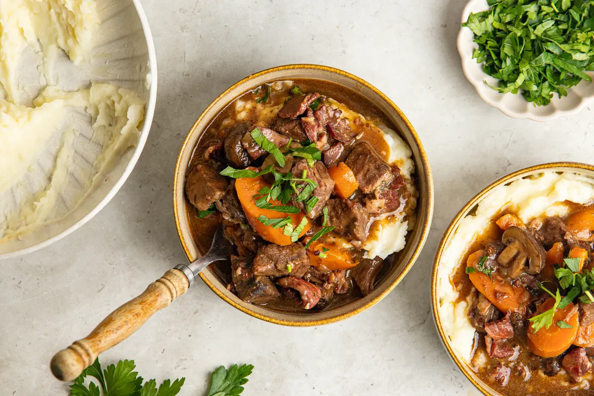 An overhead view of beef bourguignon spooned generously over creamy mashed potato, showing the tender slow-cooked beef, carrots, mushrooms and bacon in a rich sauce.