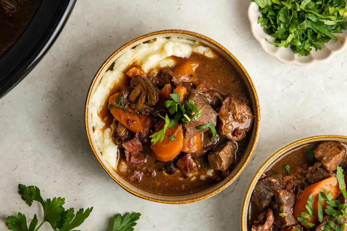 A close-up of slow cooker beef bourguignon served with fluffy mashed potato, with large pieces of tender beef, carrots, mushrooms and smoky bacon in a deep red wine gravy.