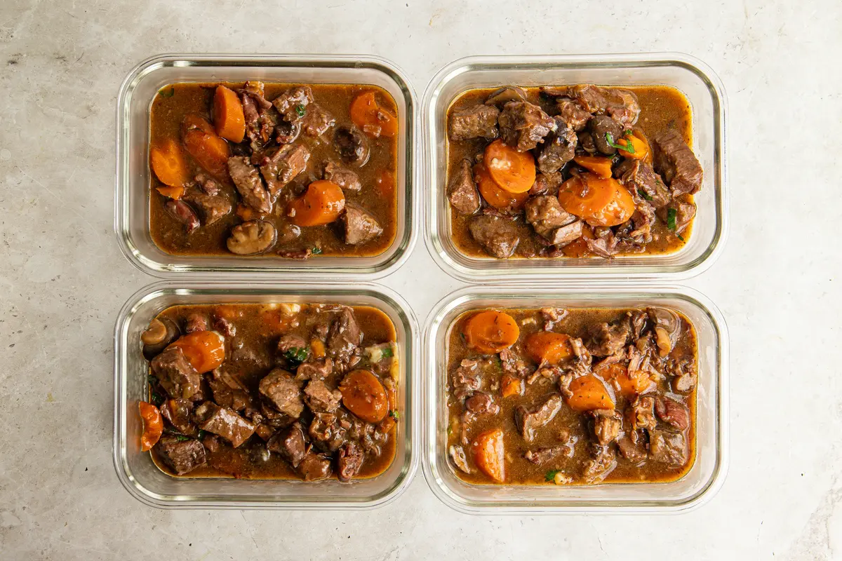 Four portions of slow cooker beef bourguignon divided into glass meal-prep containers, packed with beef, carrots, mushrooms and bacon in a rich sauce – ideal for preparing dinners ahead.