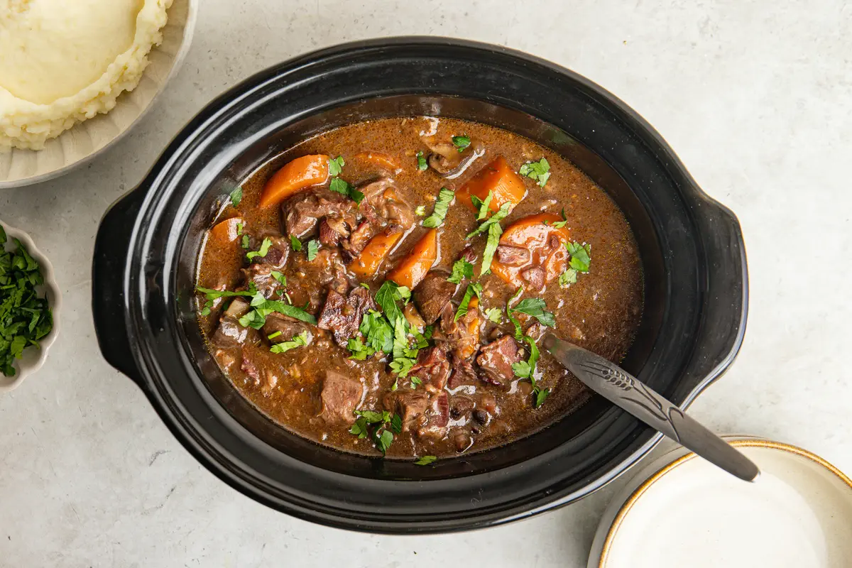 An overhead view of the finished slow cooker beef bourguignon, packed with tender beef, carrots, mushrooms and bacon in a rich red wine sauce and scattered with fresh parsley.