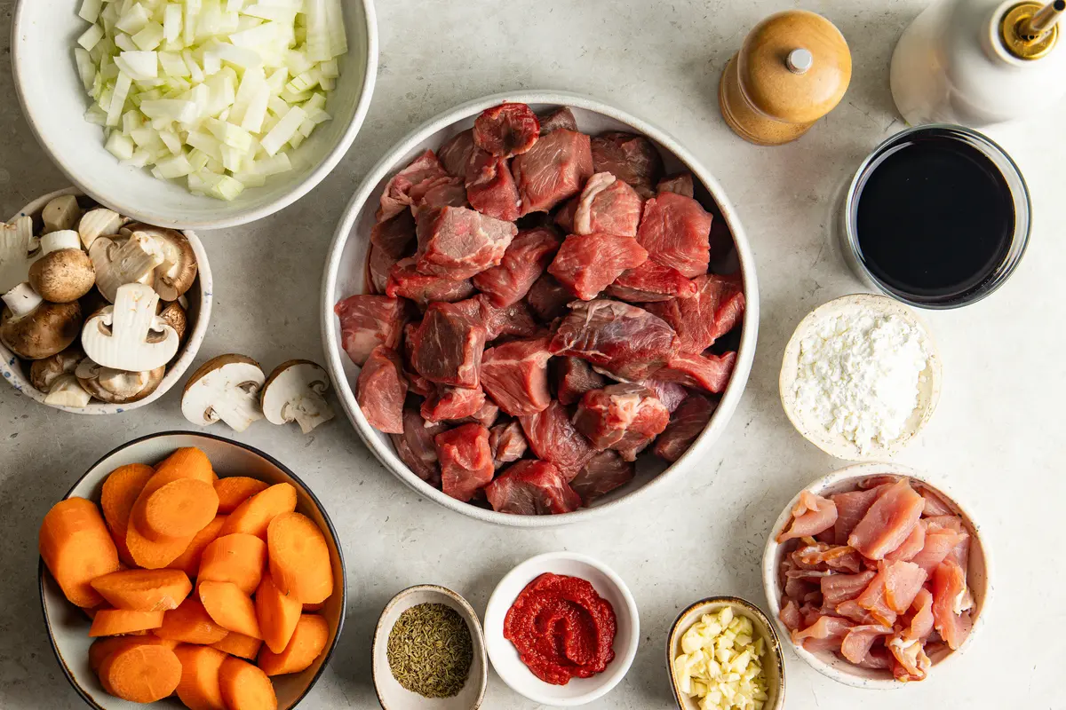 The ingredients for slow cooker beef bourguignon laid out ready for cooking, including cubed stewing beef, smoked bacon, carrots, mushrooms, onions, garlic, red wine, tomato purée, cornflour and thyme.