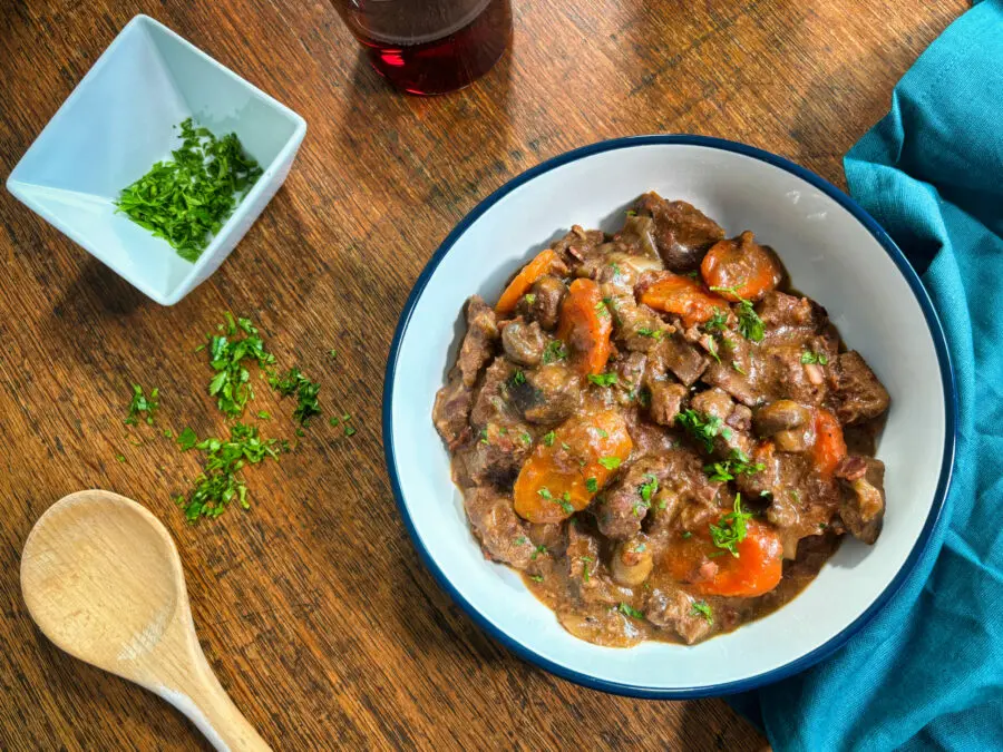 A hearty beef stew with carrots and mushrooms, garnished with fresh herbs, alongside a wooden spoon and a glass of red beverage.