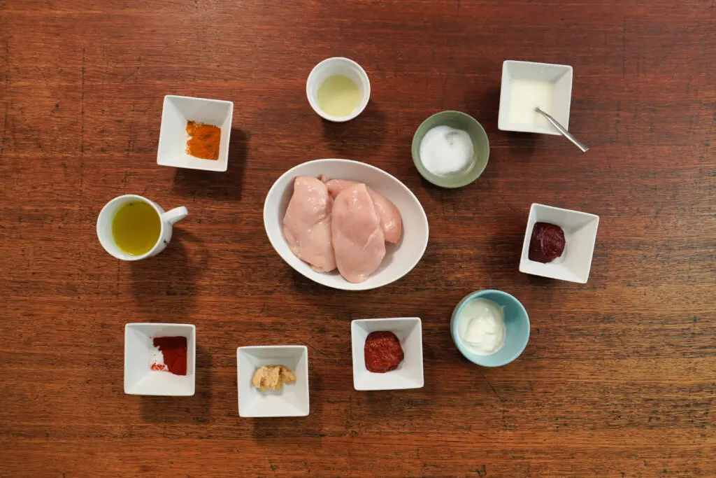 A top-down view of raw chicken breast, harissa paste, garlic, honey, lemon, smoked paprika, cumin, tomato purée, chicken stock, cornflour, salt, and Greek yoghurt, arranged neatly on a kitchen counter.