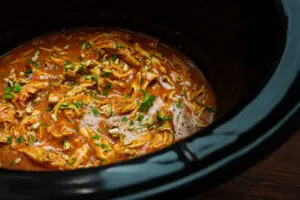 A bubbling pot of harissa chicken in the slow cooker, with tender shredded chicken submerged in a deep red, spiced sauce. The sauce has thickened, clinging to the chicken, while steam rises, hinting at its bold, aromatic flavours