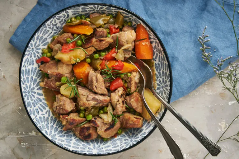 Flat lay of a bowl of slow-cooked Caldereta de Cordero, filled with tender lamb, potatoes, carrots, peppers, and peas, garnished with fresh rosemary.