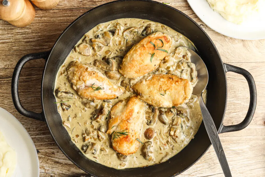 Golden chicken breasts in creamy mushroom sauce, garnished with fresh tarragon in a black pan.