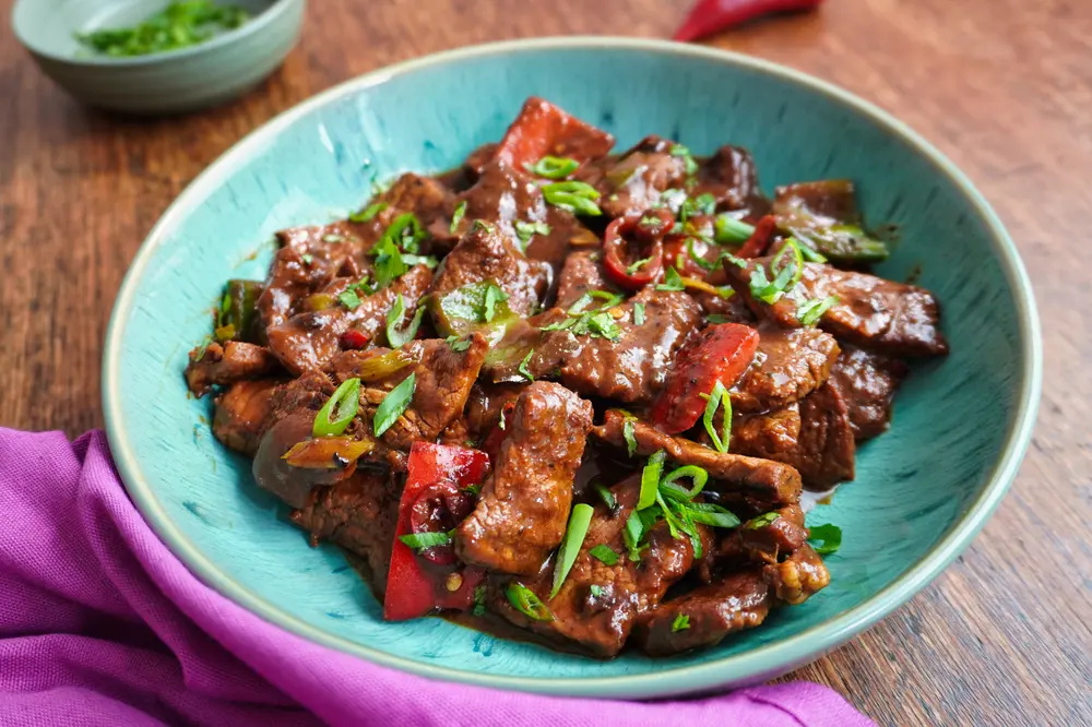 Overhead shot of Jamaican pepper steak, served in a blue bowl with a purple napkin, fresh herbs, and a red chilli in the background.