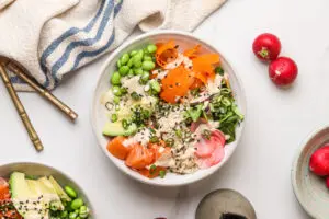 A flat lay of a salmon poke bowl arranged with fresh ingredients, including salmon, avocado, edamame, pickled radish, and shredded lettuce, all drizzled with spicy mayo and garnished with black sesame seeds.