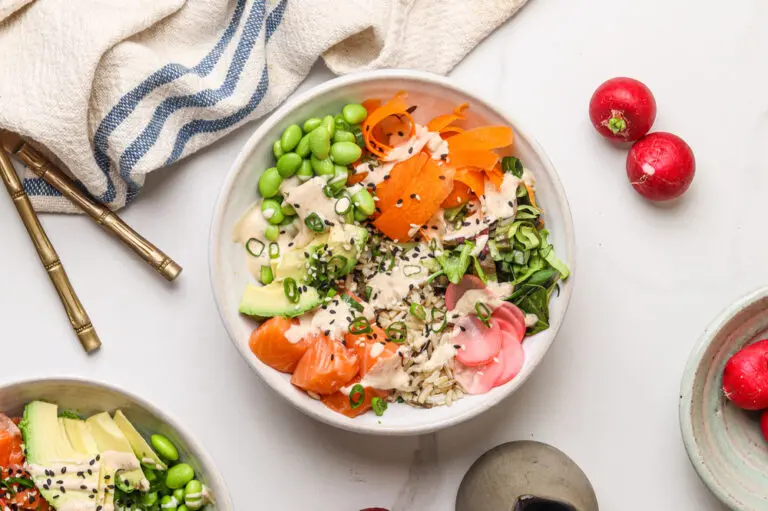 A flat lay of a salmon poke bowl arranged with fresh ingredients, including salmon, avocado, edamame, pickled radish, and shredded lettuce, all drizzled with spicy mayo and garnished with black sesame seeds.