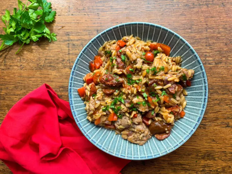 A serving of chicken and chorizo orzo in a blue ceramic bowl, garnished with fresh parsley. The dish is vibrant with chunks of chicken, slices of chorizo, red peppers, and sun-dried tomatoes, all nestled in a rich, savoury sauce. A red napkin and fresh parsley sit beside the bowl on a rustic wooden table.