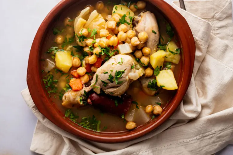 A rustic clay bowl filled with Healthy Cocido Madrileño, featuring tender chicken, chickpeas, potatoes, cabbage, and a slice of chorizo in a golden broth, garnished with fresh parsley. A soft linen napkin and silverware rest beside the bowl on a marble surface.