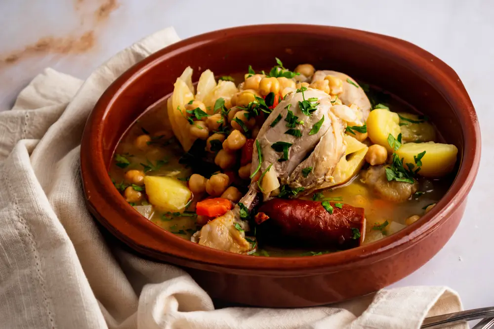 Close-up of a steaming bowl of Cocido Madrileño, showing succulent chicken thighs, plump chickpeas, chunks of potatoes, and a rich, flavourful broth with a piece of smoky chorizo. Fresh parsley adds a vibrant green contrast to the dish.