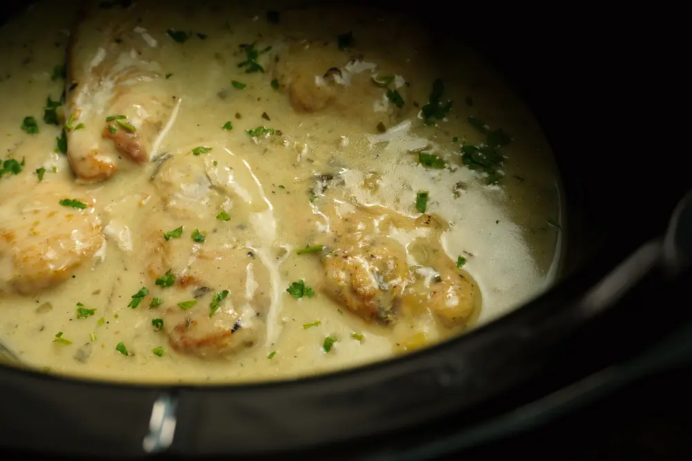 Slow cooker with creamy tarragon chicken, showcasing tender chicken pieces swimming in a thick, herby sauce, garnished with fresh parsley.