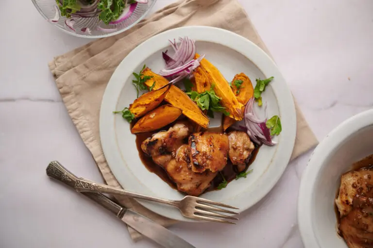 Close-up of caramelised sticky chicken thighs coated in a glossy sauce, paired with golden roasted sweet potatoes and garnished with fresh herbs.