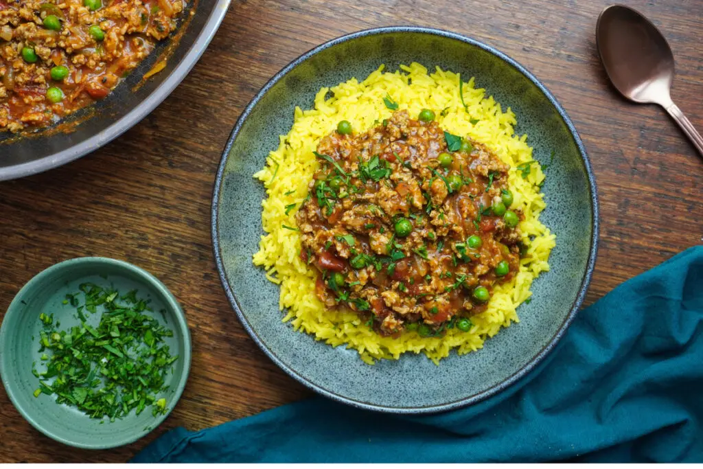 Top-down view of chicken keema served over yellow rice, garnished with fresh coriander in a ceramic bowl.