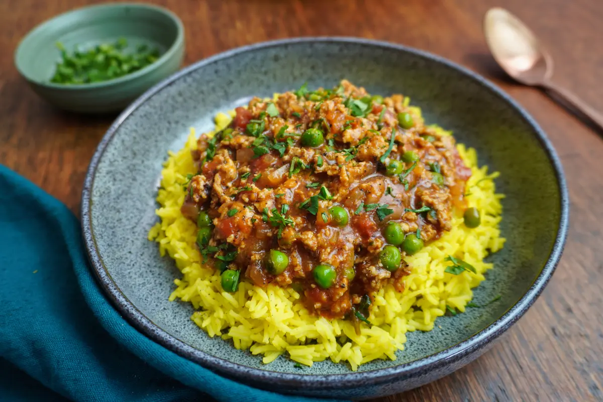 Angled shot of chicken keema and rice in a bowl, with a spoon and napkin on a wooden table.