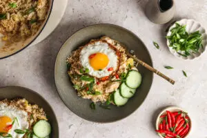 A dark bowl of golden-brown nasi goreng topped with a sunny-side-up egg, garnished with sliced red chillies, spring onions and cucumber ribbons, with extra garnish dishes alongside.