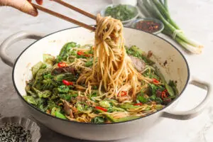 Chopsticks lifting noodles from a stir fry pan with duck and green veg