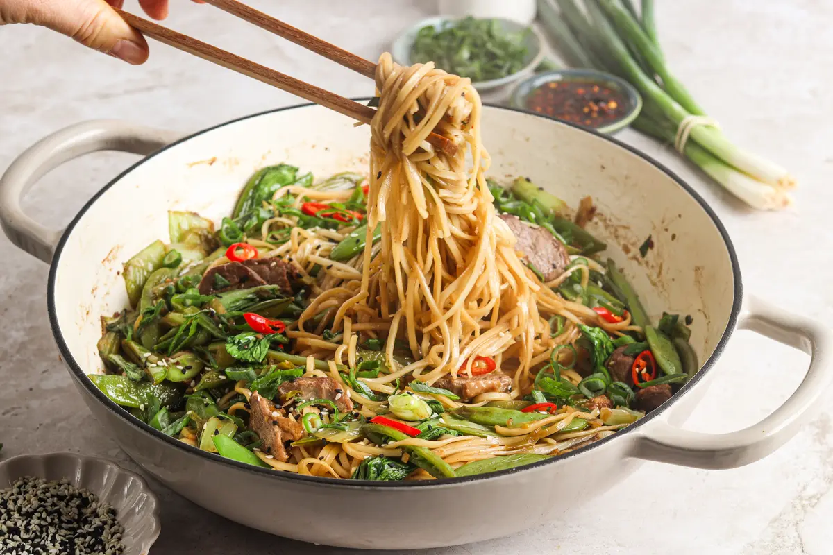 Chopsticks lifting noodles from a stir fry pan with duck and green veg