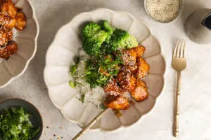 Glazed firecracker chicken with broccoli and rice in a cream bowl, topped with sesame and spring onion
