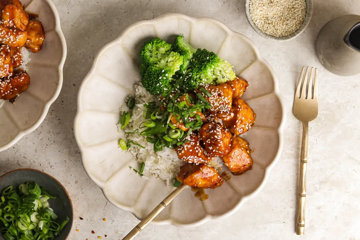 Glazed firecracker chicken with broccoli and rice in a cream bowl, topped with sesame and spring onion