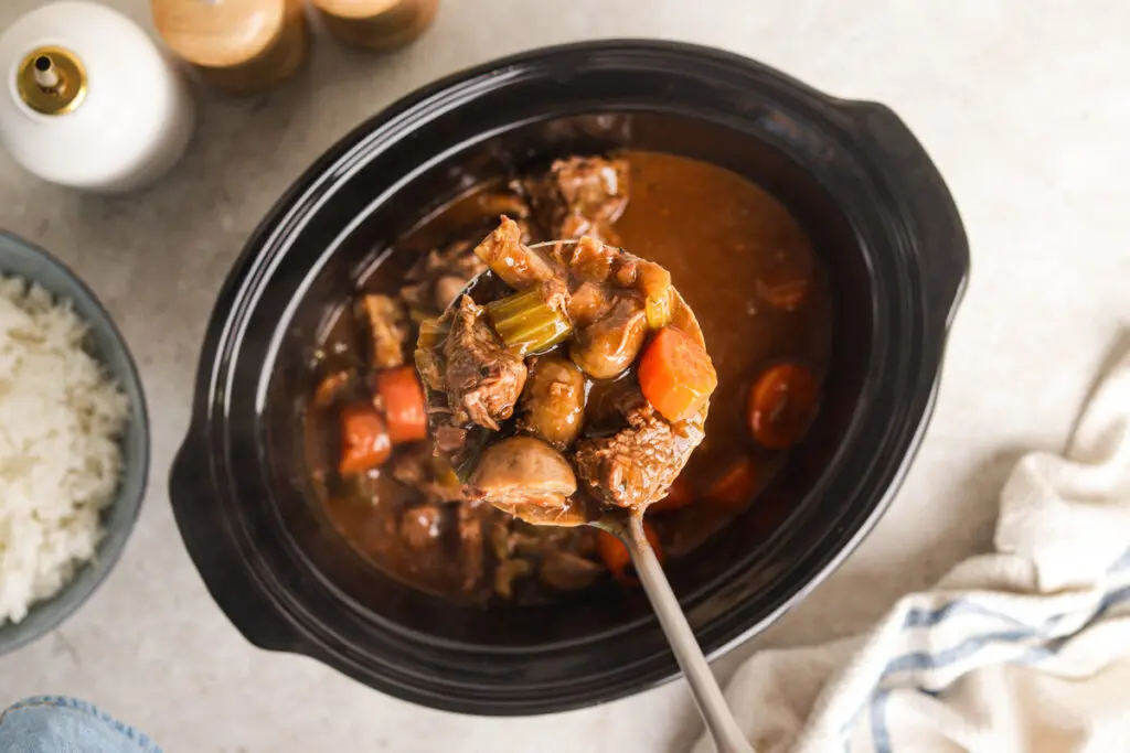 Overhead view of ladle scooping Slow Cooker Beef Casserole from pot with carrots and mushrooms.