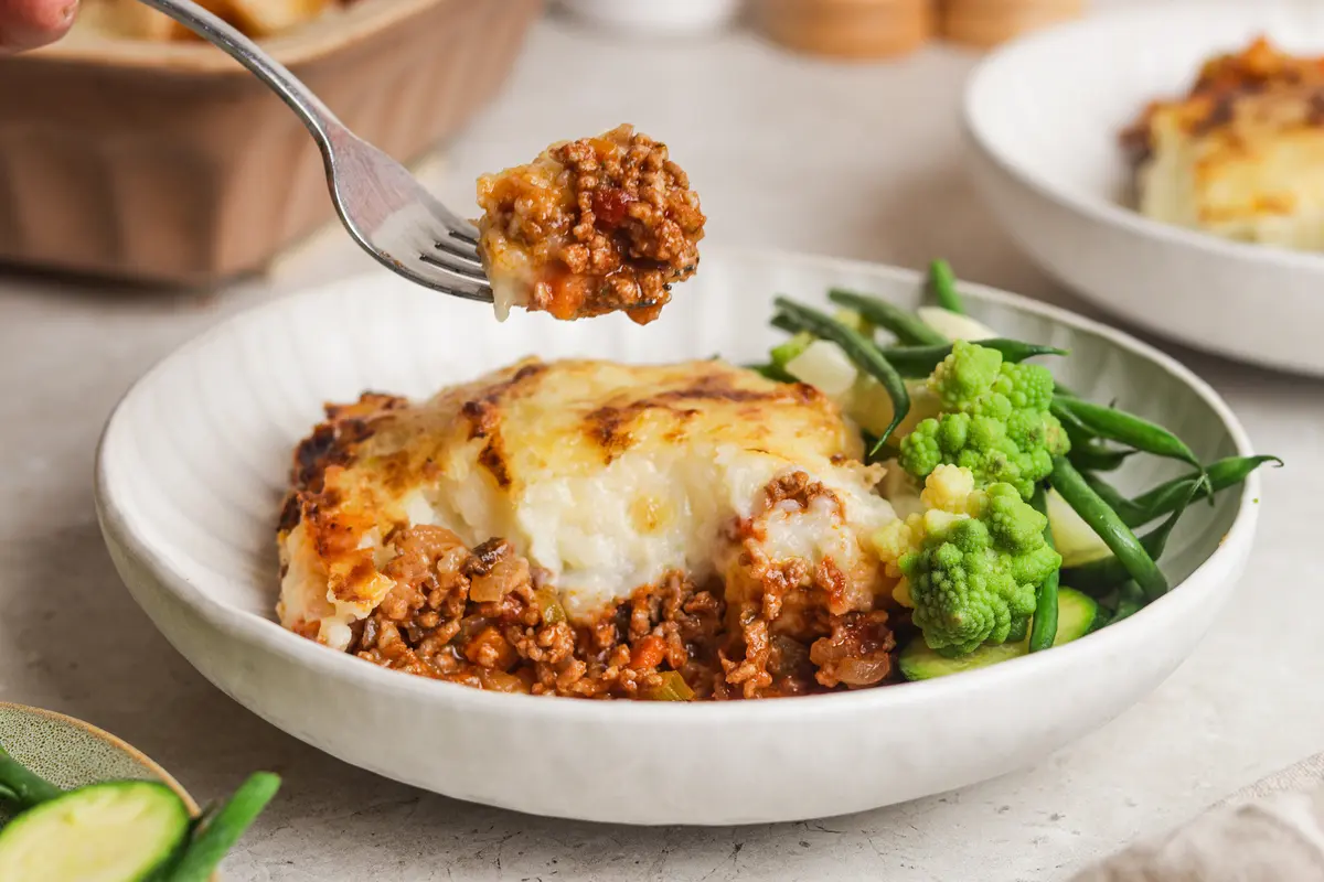 Fork lifting saucy beef mince from a plated cottage pie with vegetables
