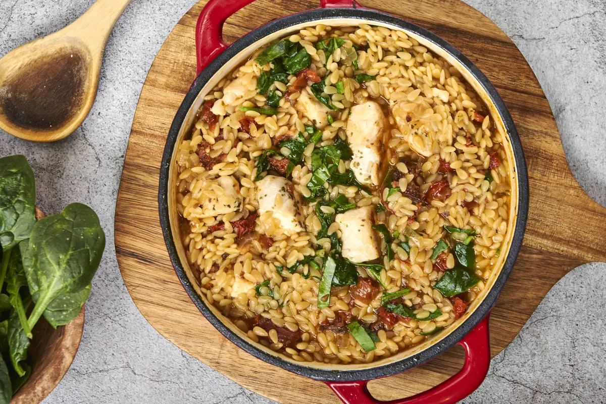 Top-down view of chicken and chorizo orzo cooked in a red pan with spinach and sun-dried tomatoes.
