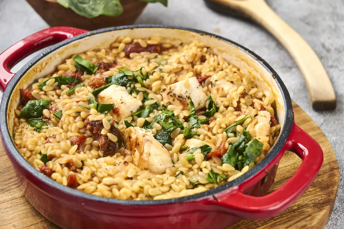 Side angle of creamy chicken and chorizo orzo in a red casserole dish with glossy tomato sauce.