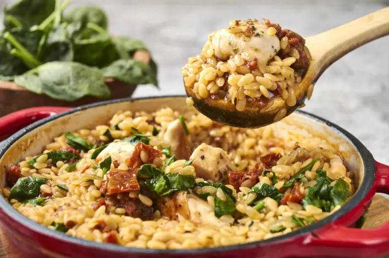 Spoon lifting chicken and chorizo orzo from the pan, highlighting creamy texture and chunky chicken.