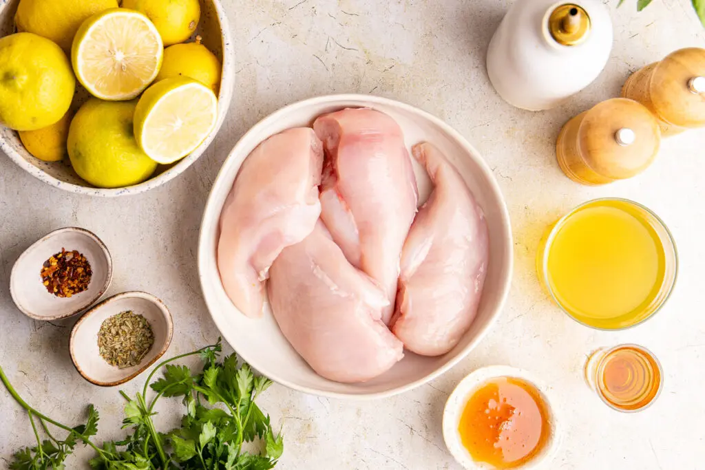Raw chicken breasts with lemon, honey, herbs, chilli flakes and stock on light surface