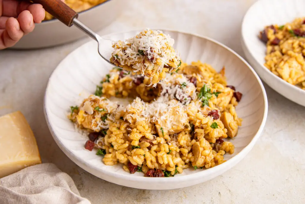 Spoon lifting creamy chicken and chorizo risotto topped with parmesan cheese from a shallow serving bowl.