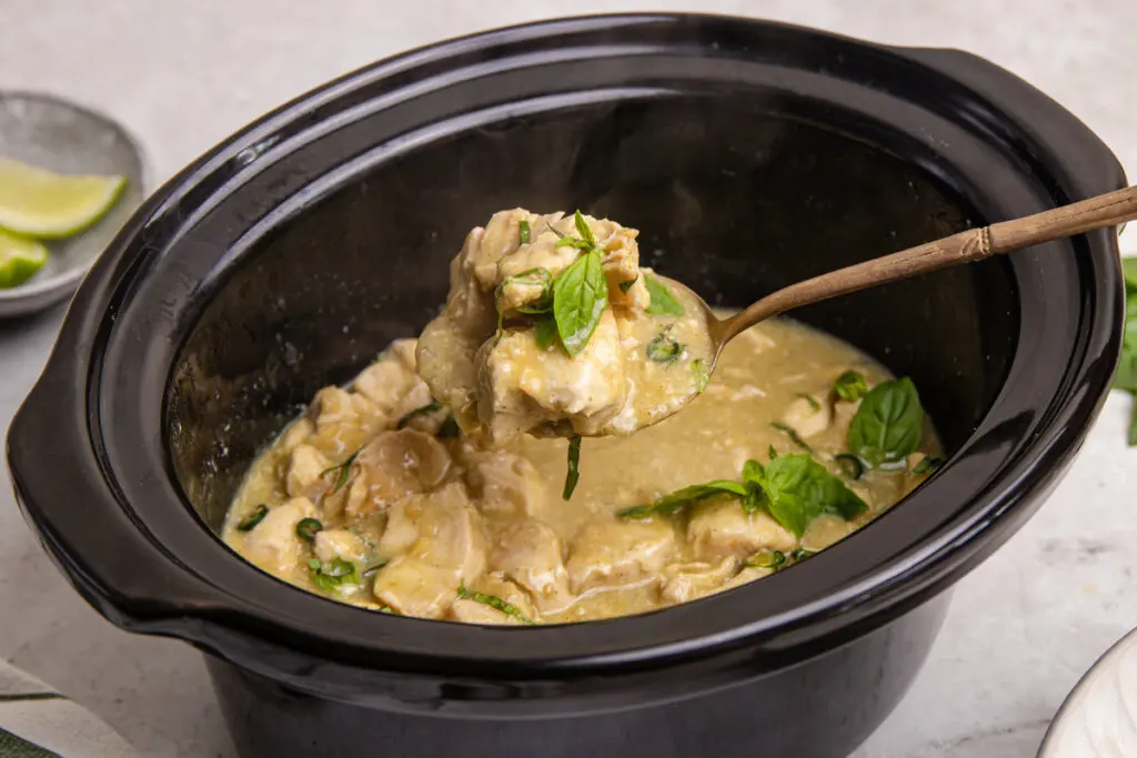 Spoon lifting tender chicken from slow cooker Thai green curry with basil garnish